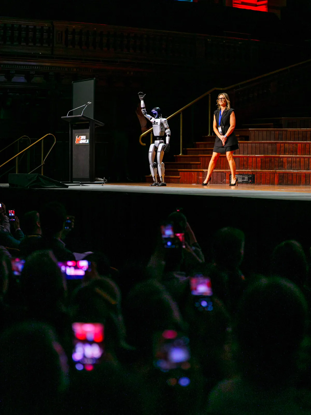 Rocco the humanoid robot waving on stage beside a presenter at OpenSummit.AI Sydney, with audience members filming
