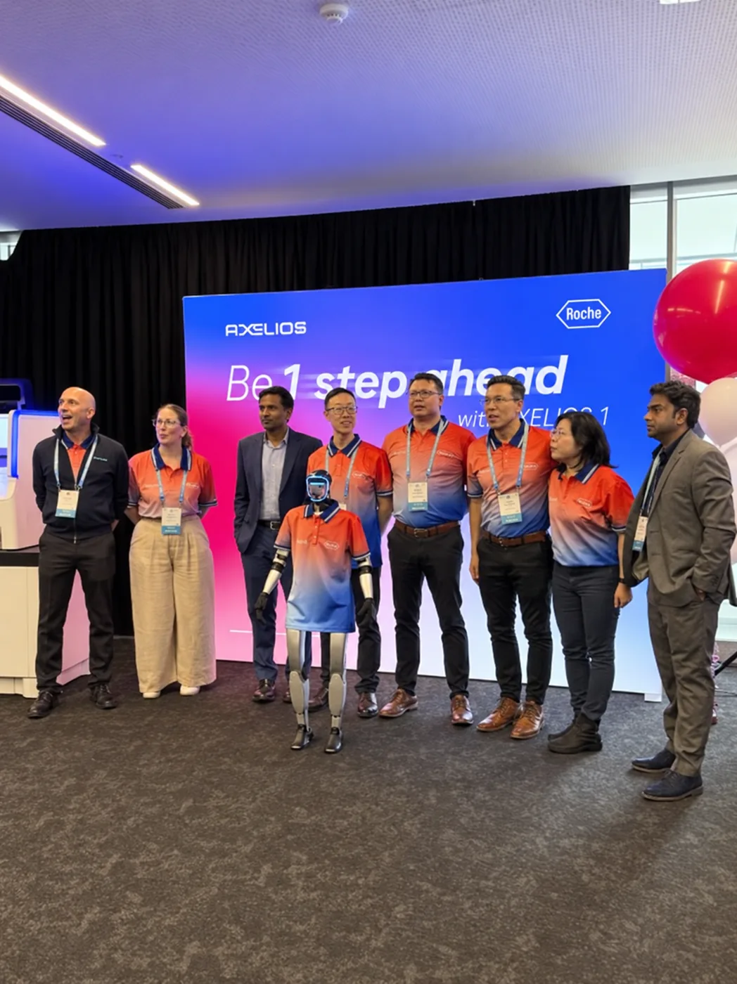 Rocco the humanoid robot in a Roche-branded polo with the Roche team in front of an AXELIOS 1 backdrop