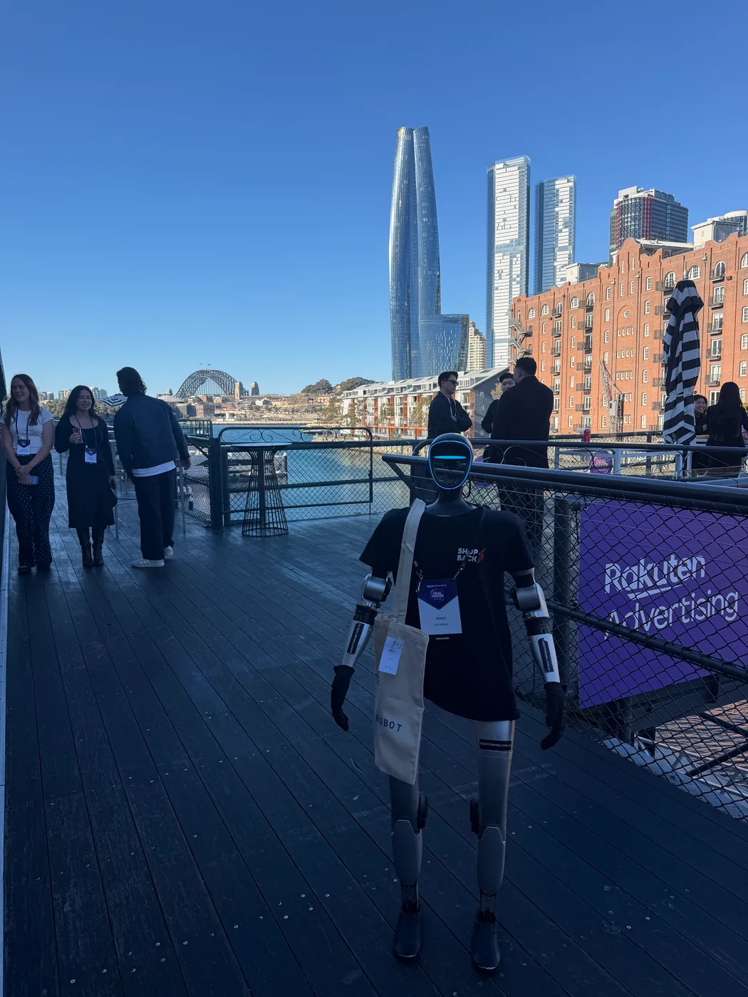Rocco the humanoid robot in a ShopBack t-shirt beside Rakuten Advertising signage at Jones Bay Wharf, with the Sydney Harbour Bridge behind